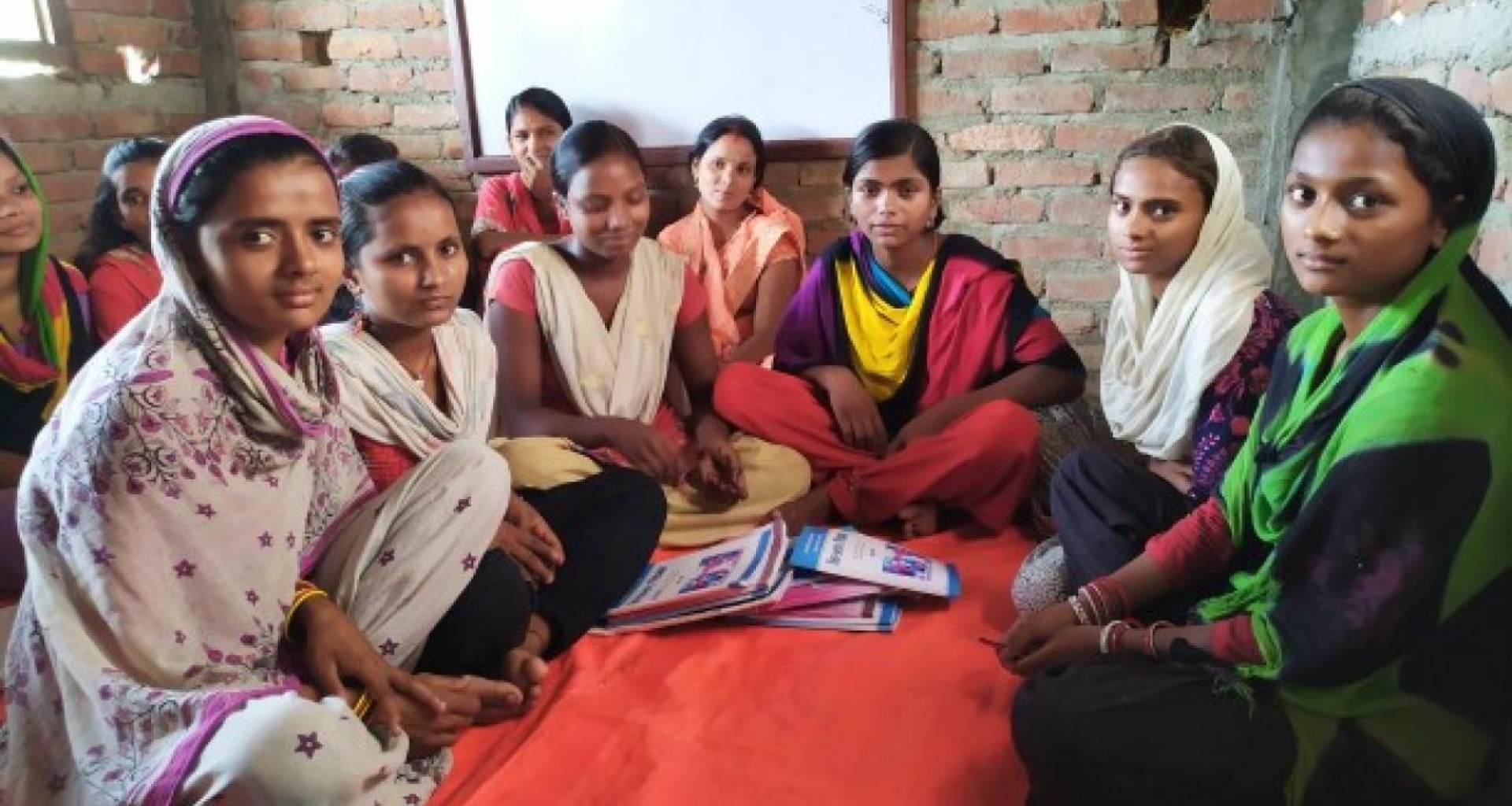A group of eleven women wearing kurthas are sitting on a red mattress inside a newly built room. There are notebooks in front of them and a whiteboard behind at the wall.